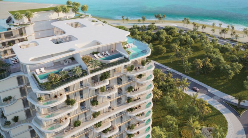 Beach Walk Grand 2 est un immeuble d'appartements moderne et luxueux en bord de mer, avec des balcons incurvés, des piscines sur le toit et une végétation luxuriante donnant sur une plage tropicale et un océan turquoise, entourés de palmiers et de jardins paysagers.