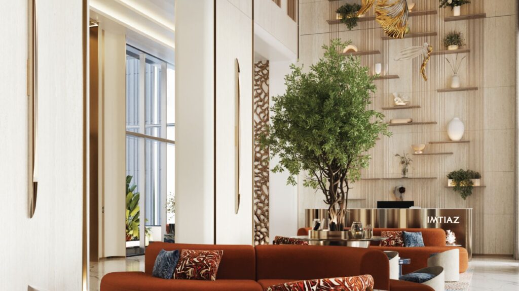 Un espace de vie moderne et haut de plafond à Beach Walk Grand comprend des canapés orange, des coussins à motifs, un grand arbre d'intérieur et des étagères décoratives avec des œuvres d'art et des plantes sur un mur crème. De grandes fenêtres inondent la pièce de lumière naturelle.