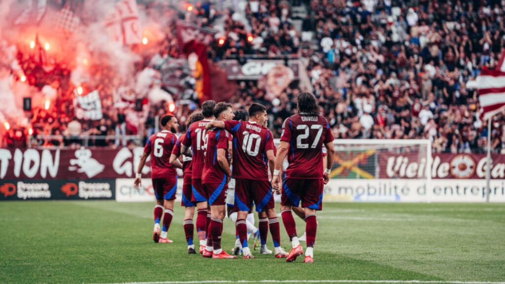 Eine Gruppe von Fußballspielern in bordeauxroten Uniformen schmiegt sich auf dem Spielfeld aneinander, unterstützt von einer Menge begeisterter Fans und leuchtend roten Leuchtraketen, und feiert Dubai Immo wird offizieller Partner des FC Servette.