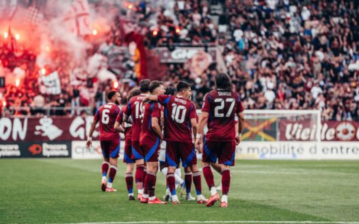 Eine Gruppe von Fußballspielern in bordeauxroten Uniformen schmiegt sich auf dem Spielfeld aneinander, unterstützt von einer Menge begeisterter Fans und leuchtend roten Leuchtraketen, und feiert Dubai Immo wird offizieller Partner des FC Servette.