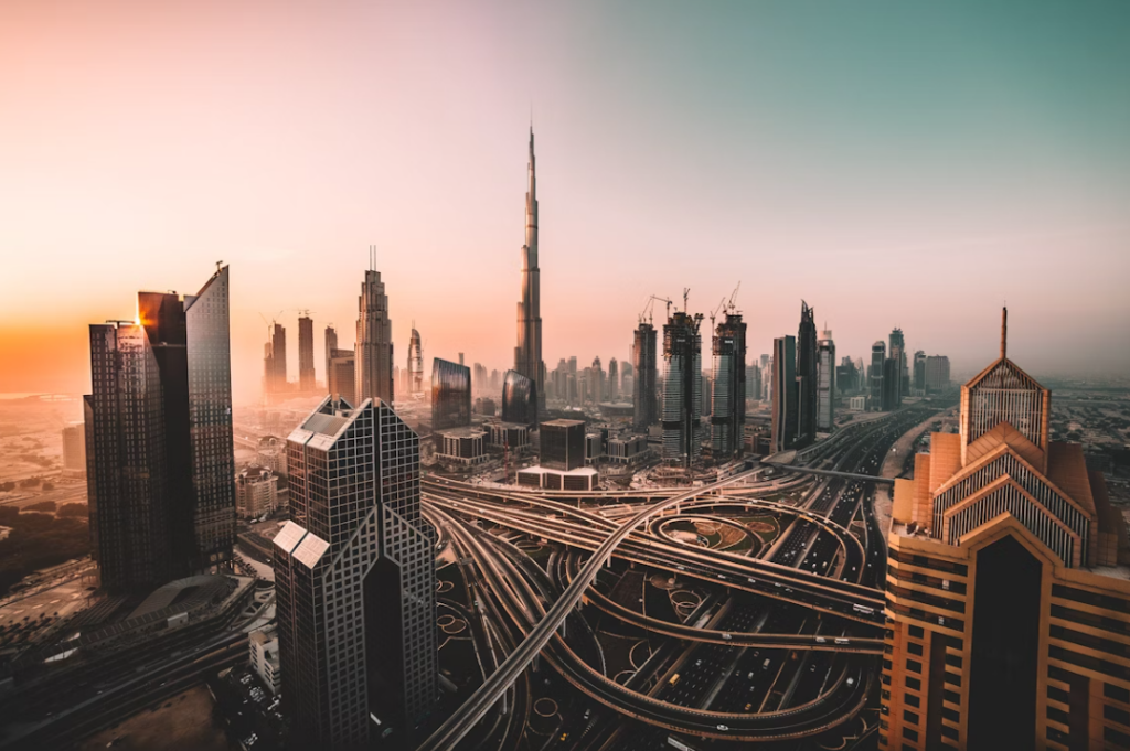 Aerial view of the Dubai skyline at sunset, with modern skyscrapers, the Burj Khalifa at the center, and an intricate network of car-filled highways under a superb gradient sky.