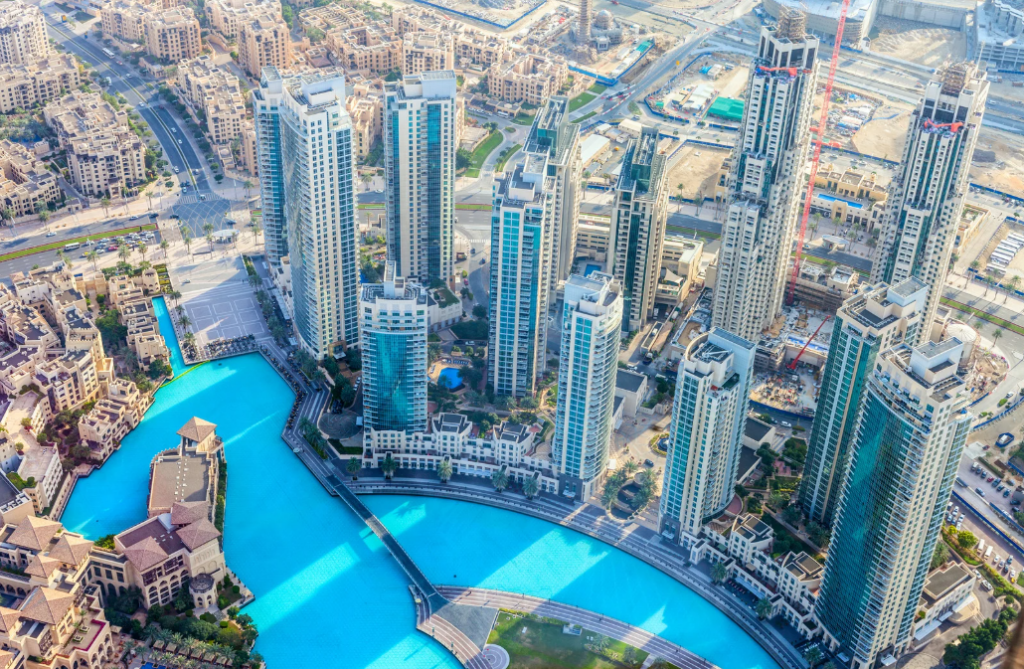 Aerial view of modern buildings surrounding a large turquoise artificial swimming pool, with roads and construction sites visible in the background, highlighting the impact of rising rents on this dynamic urban landscape.