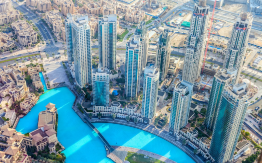 Dubai, Aerial view of modern buildings surrounding a large turquoise artificial swimming pool, with roads and construction sites visible in the background, highlighting the impact of rising rents on this dynamic urban landscape.