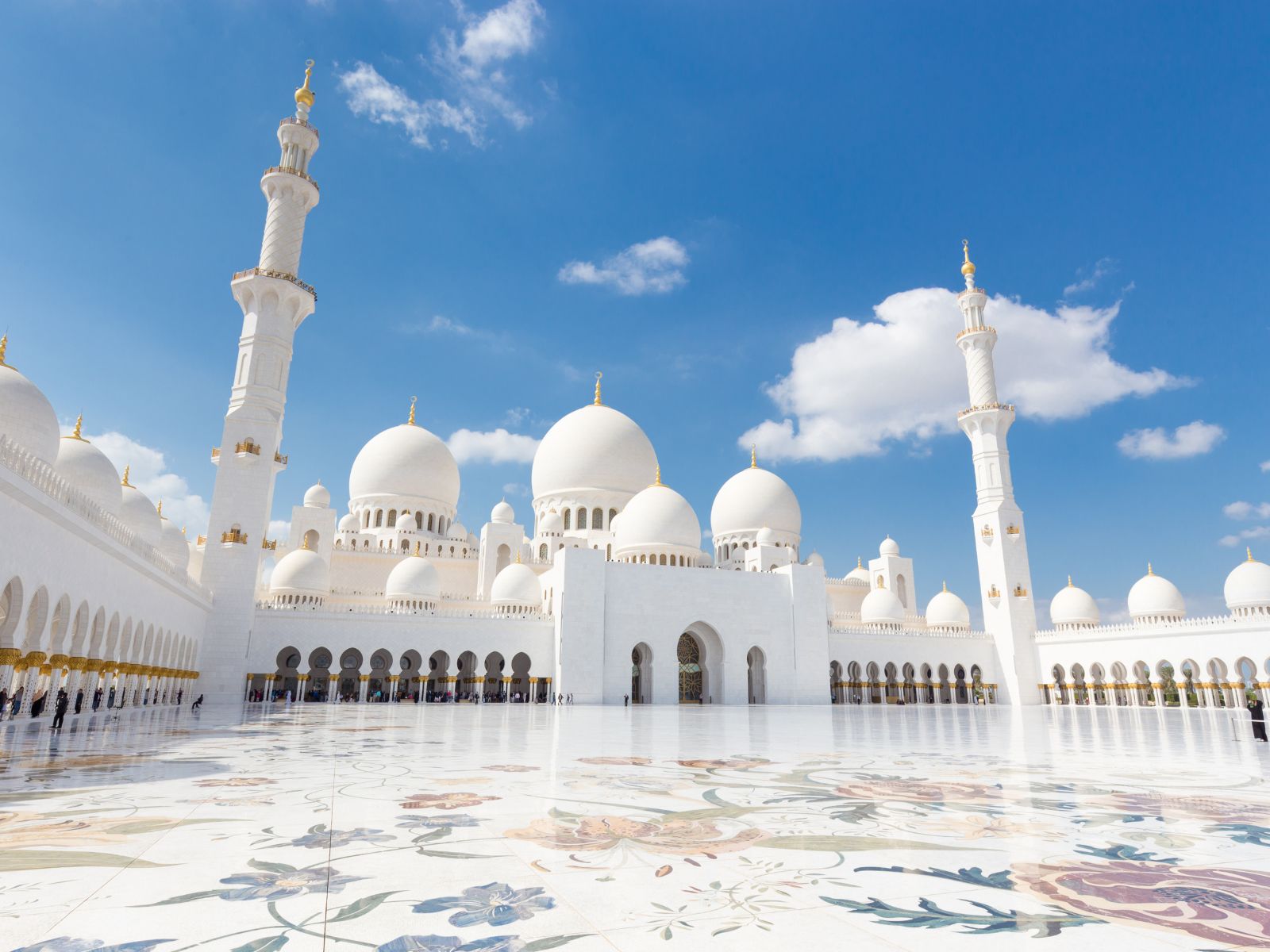 Una gran mezquita blanca con cúpulas y altos minaretes, adornada con toques dorados, se alza sobre un brillante cielo azul. El espacioso patio presenta intrincados motivos florales, una vista inspiradora para quienes buscan invertir en inmuebles en Dubai sin riba.