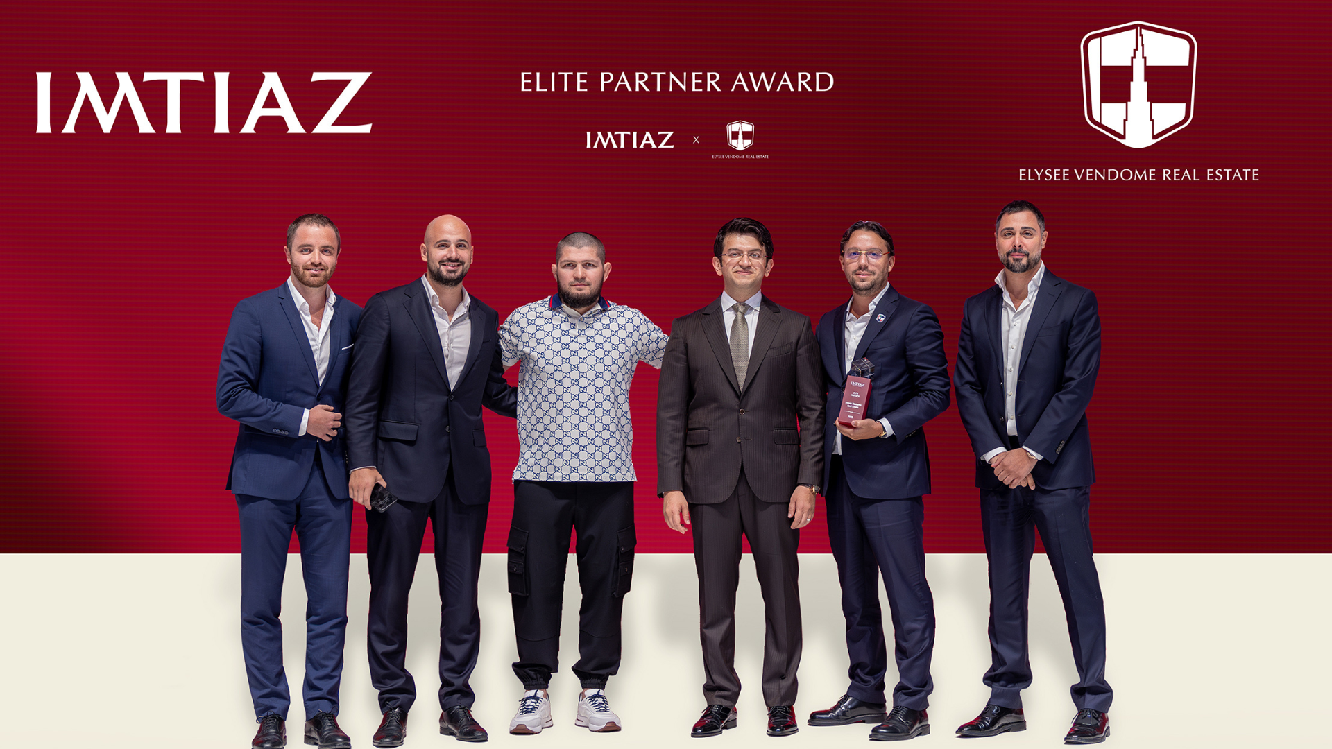 Sei uomini in abito da sera posano per una foto di gruppo davanti a uno sfondo rosso di Dubai Immo con i loghi "IMTIAZ", "Elite Partner Award" e "Elysee Vendome Real Estate". Un uomo regge con orgoglio un premio.