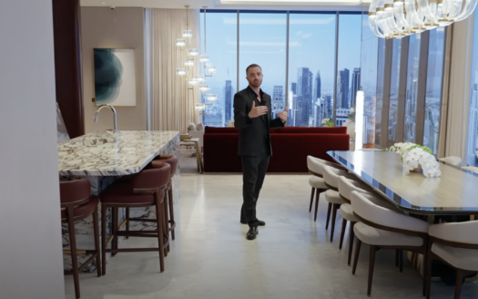 Ein Mann im Anzug steht in einem modernen, luxuriösen Apartment im Waldorf Astoria Dubai, mit Marmortheken, einem großen Esstisch und Fenstern, die vom Boden bis zur Decke reichen und auf eine atemberaubende Skyline blicken.