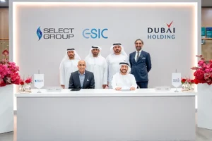 Sechs Männer in Arbeitskleidung posieren für ein offizielles Foto während einer Zeremonie zur Unterzeichnung einer Vereinbarung zwischen der Select Group und der Dubai Holding. Hinter ihnen sind die Logos der beiden Unternehmen und der ESIC zu sehen, und zu beiden Seiten des weißen Schreibtisches sind Blumenarrangements aufgestellt.