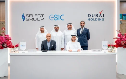 Sechs Männer in Arbeitskleidung posieren für ein offizielles Foto während einer Zeremonie zur Unterzeichnung einer Vereinbarung zwischen der Select Group und der Dubai Holding. Hinter ihnen sind die Logos der beiden Unternehmen und der ESIC zu sehen, und zu beiden Seiten des weißen Schreibtisches sind Blumenarrangements aufgestellt.