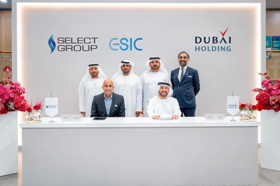 Six hommes en tenue de travail posent pour une photo officielle lors d'une cérémonie de signature d'un accord entre Select Group et Dubai Holding. Les logos des deux entreprises et de l'ESIC sont affichés derrière eux, et des compositions florales sont disposées de part et d'autre du bureau blanc.