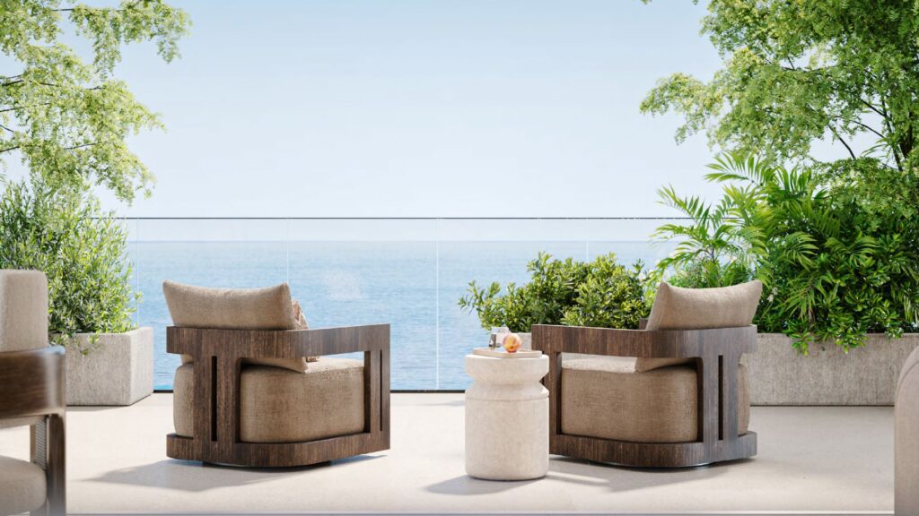 Due poltrone in legno con cuscini beige si trovano di fronte a un tavolino bianco su una terrazza che si affaccia sull'oceano a Talea, circondata da piante e alberi verdi sotto un cielo azzurro e limpido.