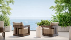 Zwei Holzsessel mit beigen Kissen stehen einem kleinen weißen Tisch gegenüber auf einer Terrasse mit Blick auf den Ozean in Talea by beyond, umgeben von grünen Pflanzen und Bäumen unter einem hellblauen Himmel.