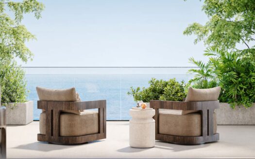 Zwei Holzsessel mit beigen Kissen stehen einem kleinen weißen Tisch gegenüber auf einer Terrasse mit Blick auf den Ozean in Talea by beyond, umgeben von grünen Pflanzen und Bäumen unter einem hellblauen Himmel.