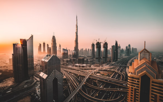Dubai cityscape at sunset, with the Burj Khalifa and modern skyscrapers. Complex highways and roads weave through the city, with warm light on the left and cooler tones on the right.