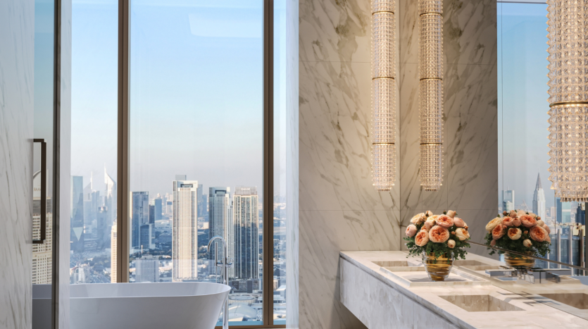 Il lussuoso bagno del Waldorf Astoria Business Bay Dubai è caratterizzato da pareti in marmo, vasca freestanding, finestre a tutta altezza con vista sulla città, due lavandini, eleganti lampadari e composizioni floreali rosa sul piano di lavoro.