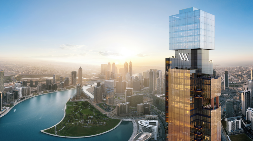 Un grande grattacielo moderno con una facciata in vetro si erge sulle rive di un fiume e si affaccia su un paesaggio urbano di parchi e grattacieli al tramonto. L'edificio presenta il logo "W" in cima.