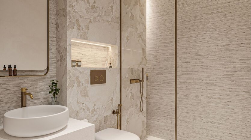 Salle de bains moderne avec des murs texturés beiges, un lavabo rond blanc, des luminaires dorés et une étagère encastrée contenant des articles de toilette belle vie zimaya, ainsi que des toilettes murales et une douche à l'italienne avec cloison en verre et pommeau de douche à effet de pluie.