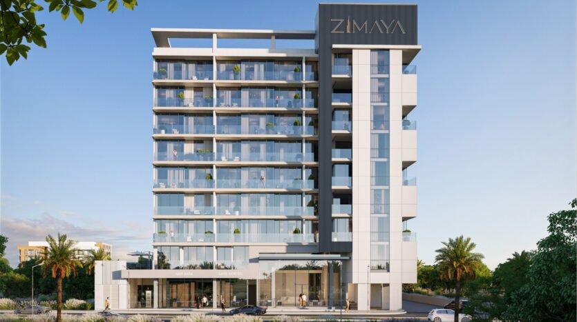 Un immeuble moderne de taille moyenne avec des balcons en verre et une enseigne "ZIMAYA" à l'angle supérieur se dresse au milieu des palmiers et de la verdure sous un ciel clair, offrant aux résidents l'expérience de la belle vie zimaya.