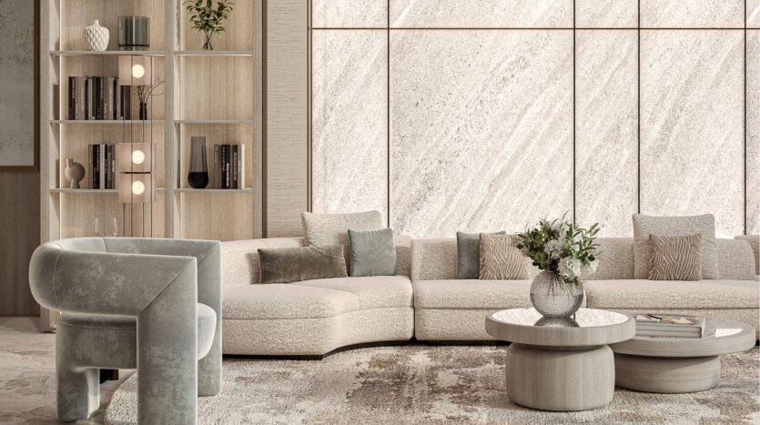Salon moderne avec un canapé sectionnel beige clair incurvé, des tables basses rondes d'inspiration belle vie zimaya, un fauteuil gris en peluche, des étagères décoratives, une grande fenêtre à la lumière naturelle et des panneaux muraux texturés. Vase avec des fleurs sur la table.