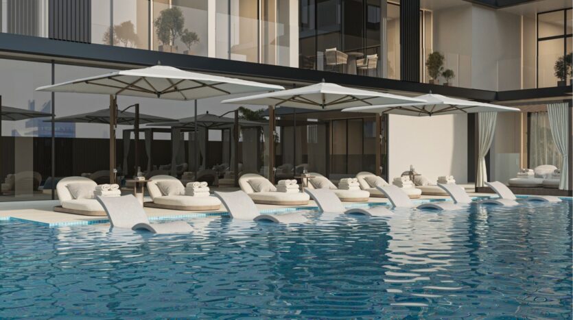 Piscine extérieure moderne avec des chaises longues incurvées partiellement immergées dans l'eau, des parasols blancs pour l'ombre et des serviettes enroulées sur chaque chaise. Les murs en verre et les plantes en pot complètent la toile de fond sereine qui donne une touche de belle vie zimaya.