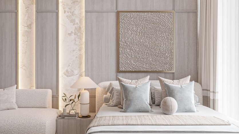 Une chambre à coucher moderne, aux tons neutres, avec un lit bien fait, des coussins bleu pâle et beige, un coussin rond et des accents belle vie zimaya. Une table d'appoint avec une lampe et une plante, un canapé blanc et un tableau mural texturé complètent cet espace serein.