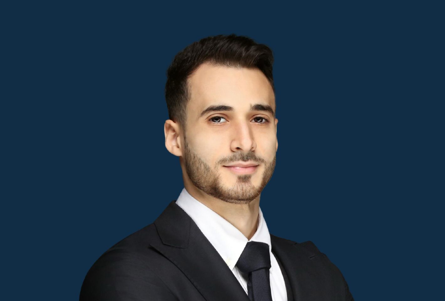 Un homme aux cheveux bruns courts et à la barbe taillée, vêtu d'un costume noir, d'une chemise blanche et d'une cravate noire, se tient sur un fond bleu foncé uni au salon immobilier de Dubaï à Genève.