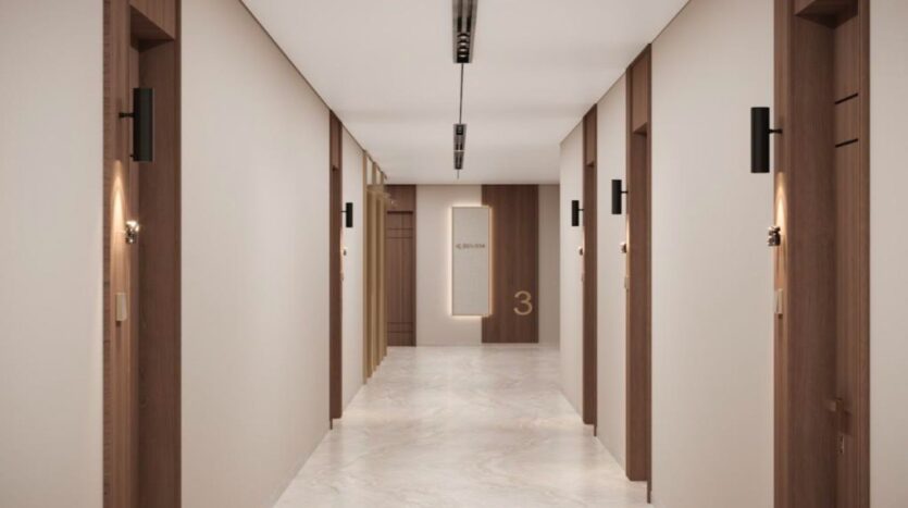 A clean, modern hallway at Marquis One Arjan features cream-colored walls, marble floors, wooden door frames and wall sconces. Doors line both sides, and a sign reading "3" is visible at the end of the hallway.