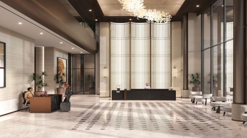 Spacious, modern lobby of the Marquis One Arjan hotel, with high ceilings, elegant lighting, central reception desk, lounge area to the left, large windows, and two people interacting near luggage.