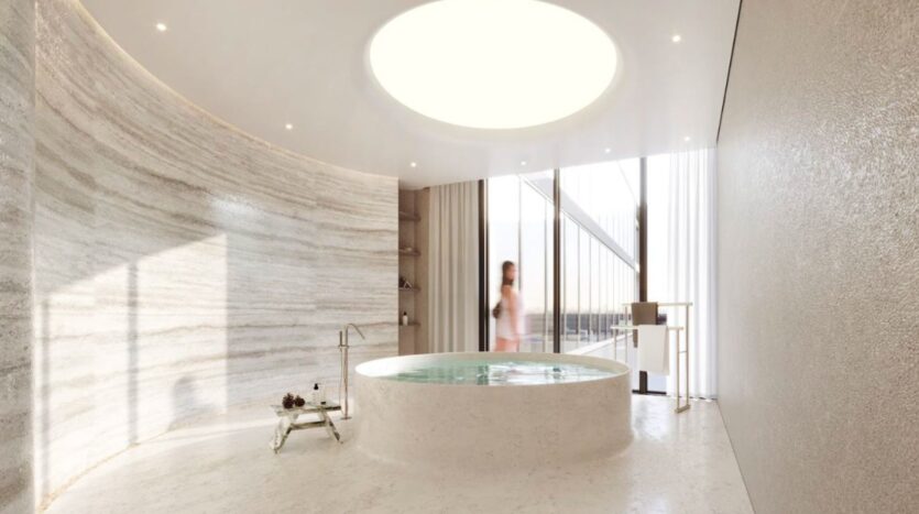 Marquis One Arjan's modern, minimalist bathroom features a large round bathtub in the center, a skylight above, curved marble walls and floor-to-ceiling windows. A scantily clad person stands by the window.