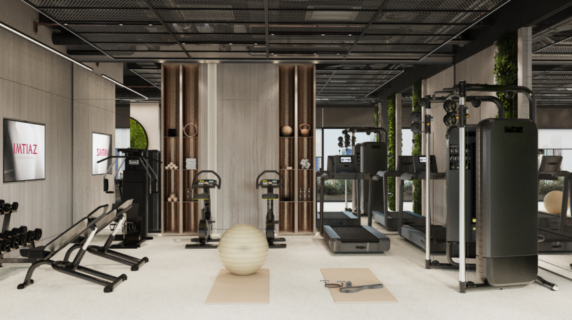 Moderno gimnasio equipado con máquinas de ejercicios, pesas libres, bancos, dos bicicletas estáticas, una pelota de estabilidad, dos colchonetas y grandes espejos. El espacio presenta tonos neutros, detalles en madera y luz natural procedente de grandes ventanales.