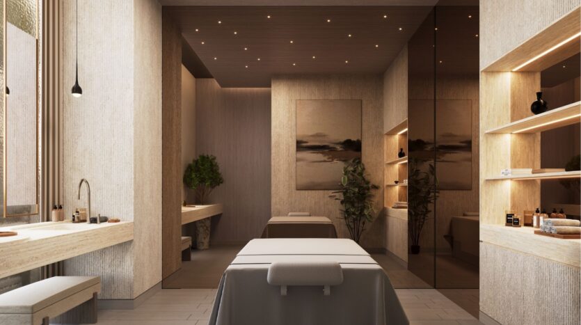 The modern spa room at Riverton House by Ellington Properties features two massage tables, soft beige lighting, wood accents, wall art, shelves with towels and products, a sink and lush plants for a calm, relaxing atmosphere.