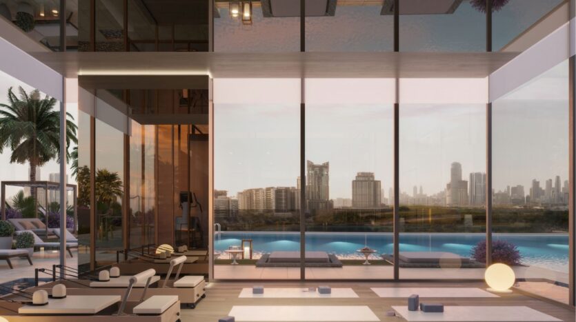 The modern fitness studio at Riverton House by Ellington Properties features floor-to-ceiling windows overlooking the pool, lounge, palm trees and city sunset. Meditation mats and equipment are neatly arranged on the wooden floor.