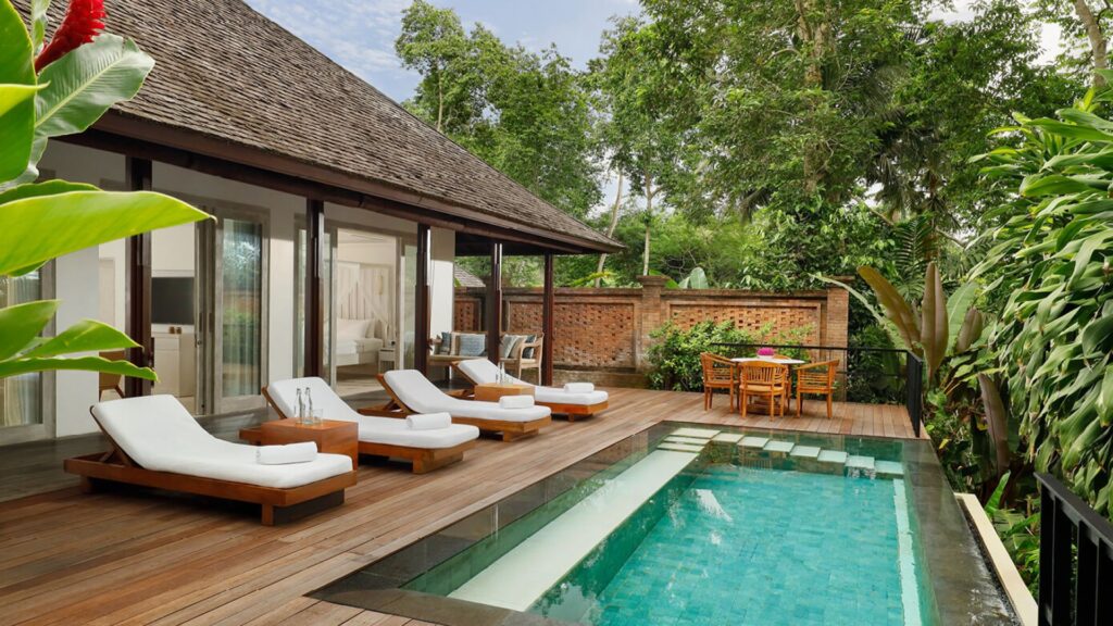 Der Innenhof einer tropischen Villa erinnert an den Luxus, den man im Vergleich zwischen Dubai, Marrakesch und Bali findet, mit drei weißen, gepolsterten Liegestühlen am Rand eines rechteckigen Pools, einem hölzernen Esstisch und üppiger Vegetation, die große Glastüren und ein Schrägdach umgibt.