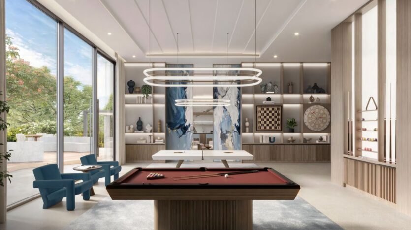 Une salle de jeux moderne à Windsor House de Ellington Properties comprend une table de billard rouge, des chaises bleues, de grandes fenêtres et des étagères encastrées avec des livres, un échiquier et un jeu de fléchettes. Des luminaires uniques et la lumière naturelle remplissent cet espace élégant.