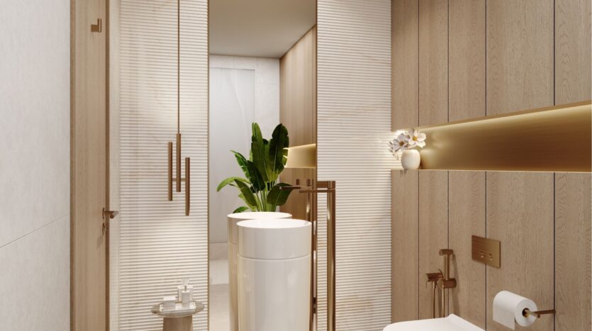 Salle de bain moderne à wynwood horizon de Imtiaz avec des murs en bois clair, un lavabo blanc, un robinet doré, des toilettes et une plante près des portes en verre dépoli. Une étagère dorée avec des objets décoratifs se trouve au-dessus du porte-papier hygiénique.
