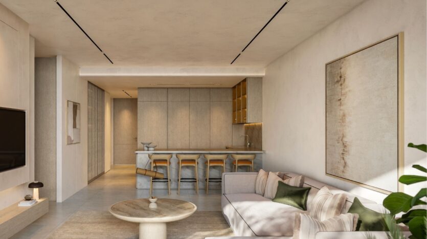 Modern living room and kitchen at Avida Residences Dubai Islands with neutral tones, a beige sofa, green cushions, a round coffee table, bar stools on a marble island, a large abstract wall painting and minimalist decor, well lit by natural and recessed lights.