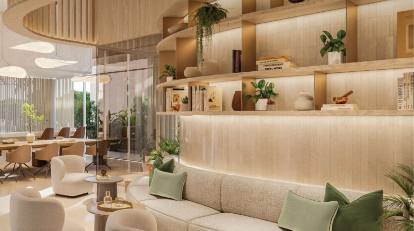 Modern living room at Avida Residences Dubai Islands with curved beige sofa, green cushions, round tables, plants, decorative shelves and large windows letting in natural light. The dining room with bronze-colored chairs is visible in the background.
