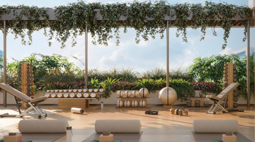 The outdoor fitness area at Avida Residences Dubai Islands features benches, dumbbells, kettlebells and yoga mats. Climbing plants are suspended from a pergola above lush plants, creating a tranquil outdoor gym atmosphere.