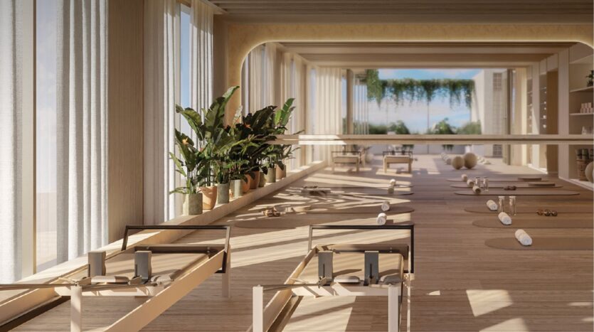 The bright, modern Pilates studio at Avida Residences Dubai Islands features large windows, wooden floors, potted plants, reformer machines, exercise mats and rolled towels. The space is bathed in sunlight and greenery is visible outside.