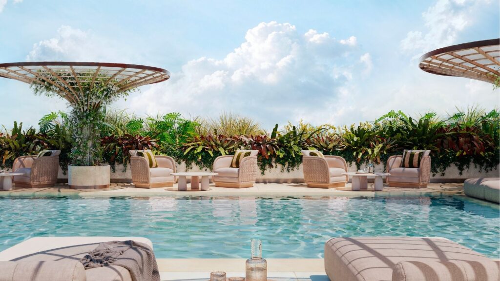 The luxurious outdoor pool at Avida Residences Dubai Islands, with sun loungers, tables and armchairs surrounded by lush tropical plants, under a bright blue sky dotted with clouds.