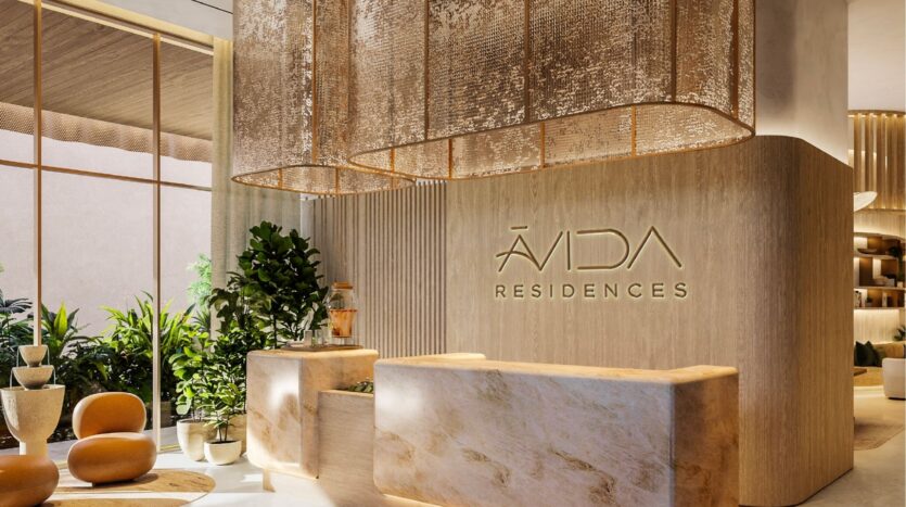 Modern, elegant hotel reception with light wood finishes, marble counters, green plants and large decorative lamps. The wall bears the inscription "ÁVIDA RESIDENCES" in gold lettering. Natural light fills the space at avida residences dubai.