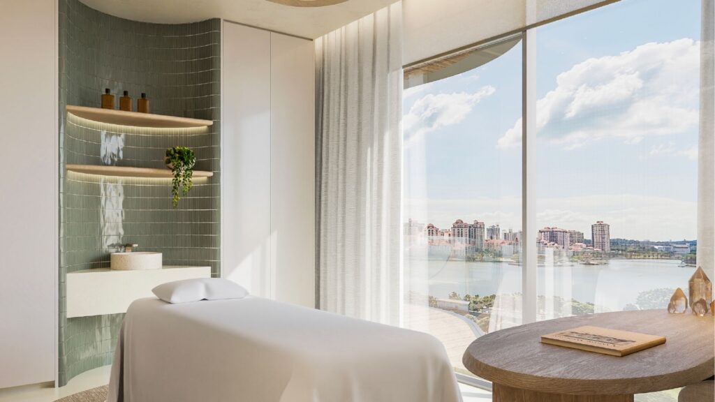 The bright, modern spa room at Avida Residences Dubai Islands features a massage table, large windows with city and water views, minimalist décor, shelves with bottles, a small green plant and abundant natural light that fills the relaxation space.