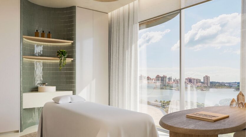 The bright, modern spa room at Avida Residences Dubai Islands features a massage table, large windows with city and water views, minimalist décor, shelves with bottles, a small green plant and abundant natural light that fills the relaxation space.