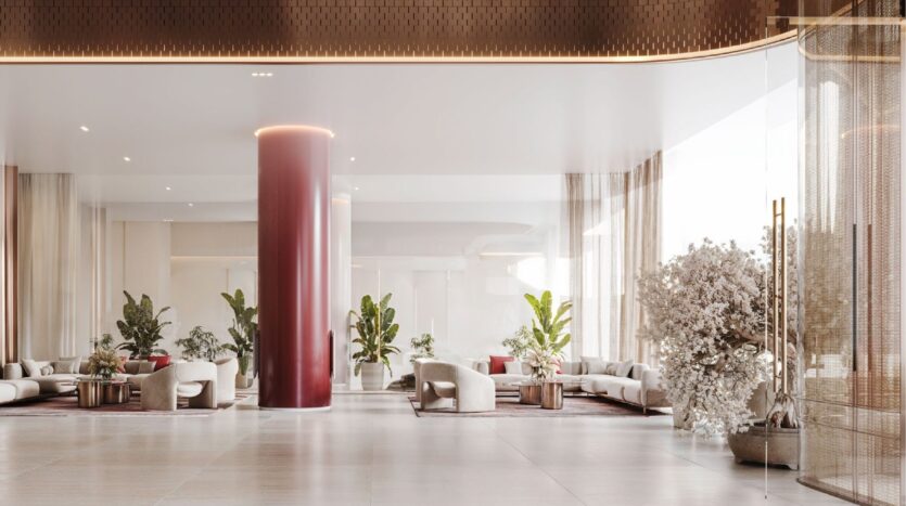 El moderno y espacioso vestíbulo del Binghatti Skyblade presenta una brillante columna roja, sofás de colores claros, plantas de interior, cortinas transparentes y grandes ventanales que inundan el espacio de luz natural. Los tonos neutros y el mobiliario elegante y minimalista completan el conjunto.