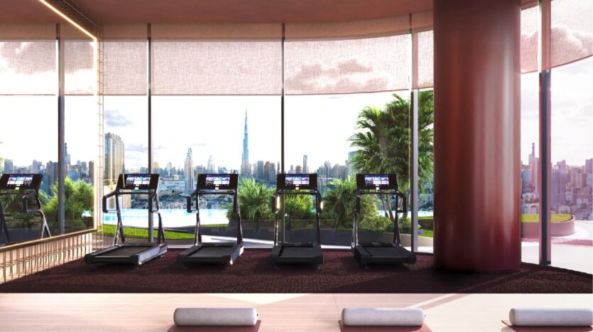 Cuatro cintas de correr dan a grandes ventanales con vistas al perfil de la ciudad y a las altas palmeras del exterior de Binghatti Skyblade. Una piscina y tumbonas esperan fuera, mientras que la luz del sol inunda este luminoso y moderno gimnasio, con esterillas de yoga en primer plano.