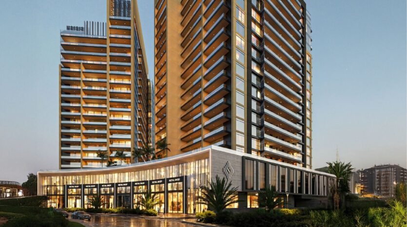 The modern Butterfly Towers Arjan, with their large balconies and bay windows, are illuminated in the evening. A low-rise shopping area with stores and palm trees lies ahead, reflecting the city lights on the wet pavement.