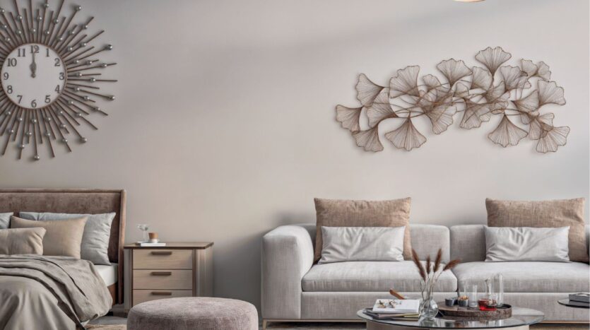 Modern living room in neutral tones in Arjan Butterfly Towers, featuring a bed, a beige sofa with cushions, a side table, a round ottoman, a decorative metal wall sculpture and a large sun-shaped wall clock.