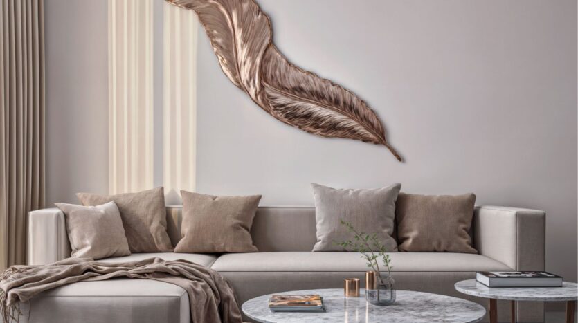 A modern living room with a beige sectional sofa, neutral cushions, a cozy blanket, round marble coffee tables with books and a plant, and metallic feather wall art on a light wall - perfect for contemporary spaces like Butterfly Towers Arjan.