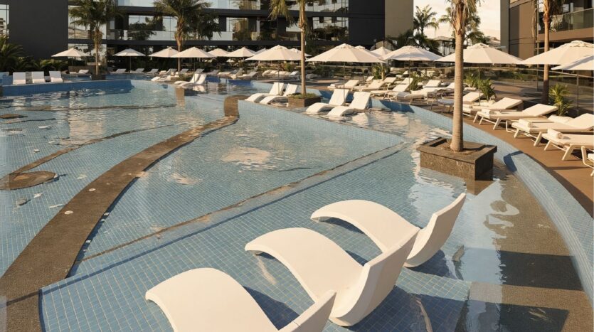 A pool with submerged white sun loungers, surrounding palm trees and numerous deckchairs shaded by large white parasols, set against the backdrop of the modern architecture of the Arjan Butterfly Towers and elegant poolside seating.