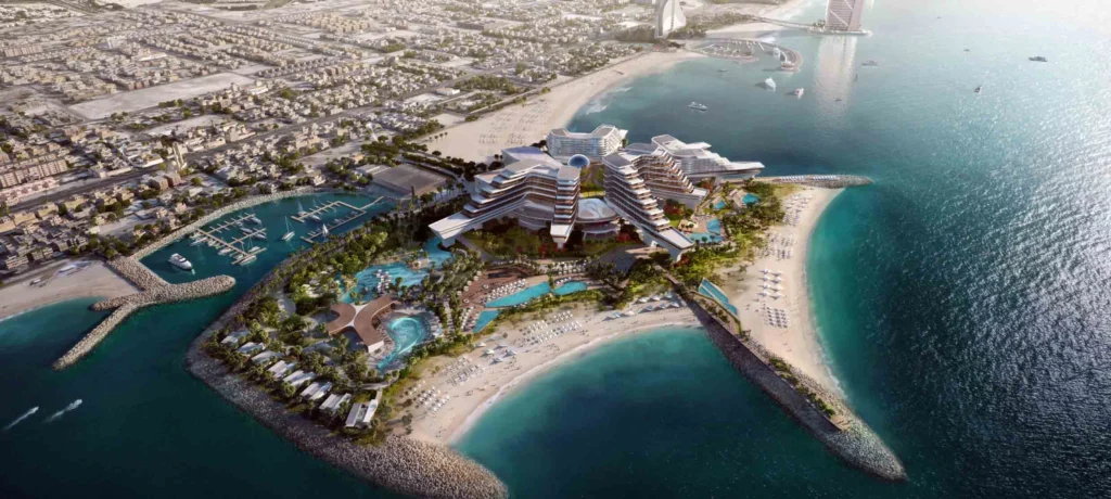 Aerial view of a modern resort on an artificial island in Dubai, with swimming pools, sandy beaches, palm trees and surrounding waters near the city's coastline and marinas, with buildings, roads and an MGM casino in the background.