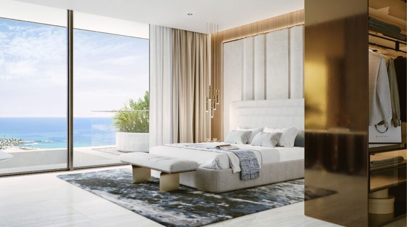Chambre à coucher moderne avec un grand lit, des tons neutres et un tapis en peluche. Des fenêtres allant du sol au plafond offrent une vue sur l'océan et les îles Silena Dubaï, tandis qu'un placard ouvert contenant des vêtements est visible sur la droite. La lumière naturelle remplit l'espace.